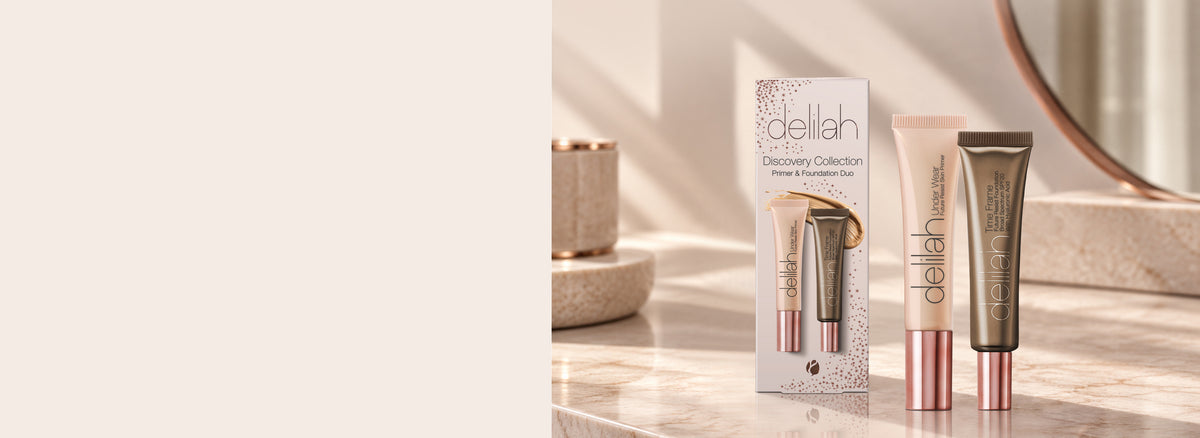 A Delilah cosmetics discovery set featuring a cream-colored tube, a gold-toned tube, and its retail box on a marble surface with a mirror in the background.